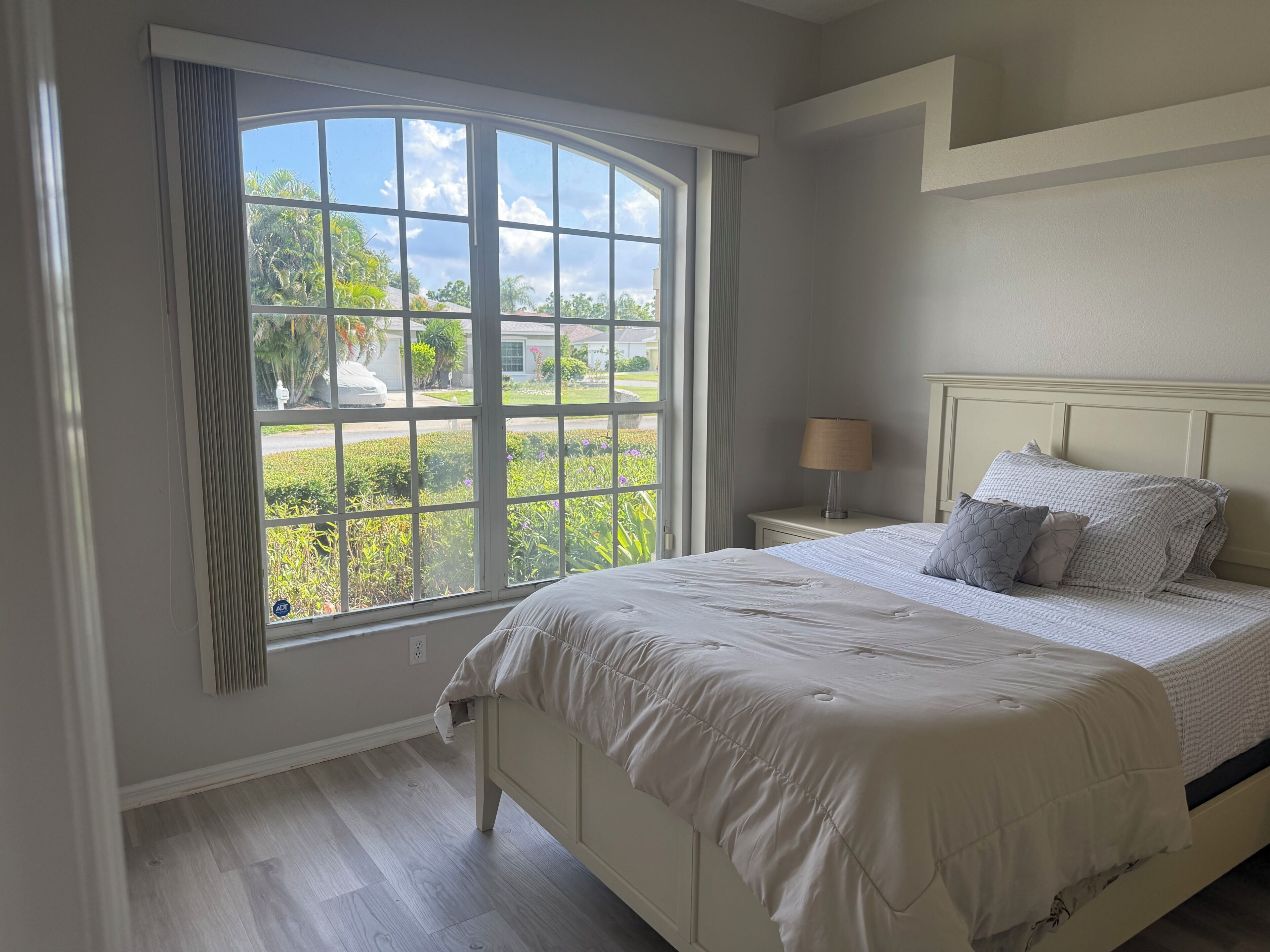 A bedroom with a large window, a bed with a white headboard, and a bedside table with a lamp.