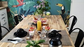Una mesa de madera con un mural tropical, un vaso de jugo, un bol de fruta, un hervidor y una pequeña planta.