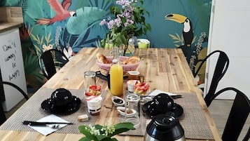 Une table de salle à manger en bois ornée d’une fresque aux motifs tropicaux, un verre de jus de fruits, un saladier rempli de fruits, une théière et une petite plante.