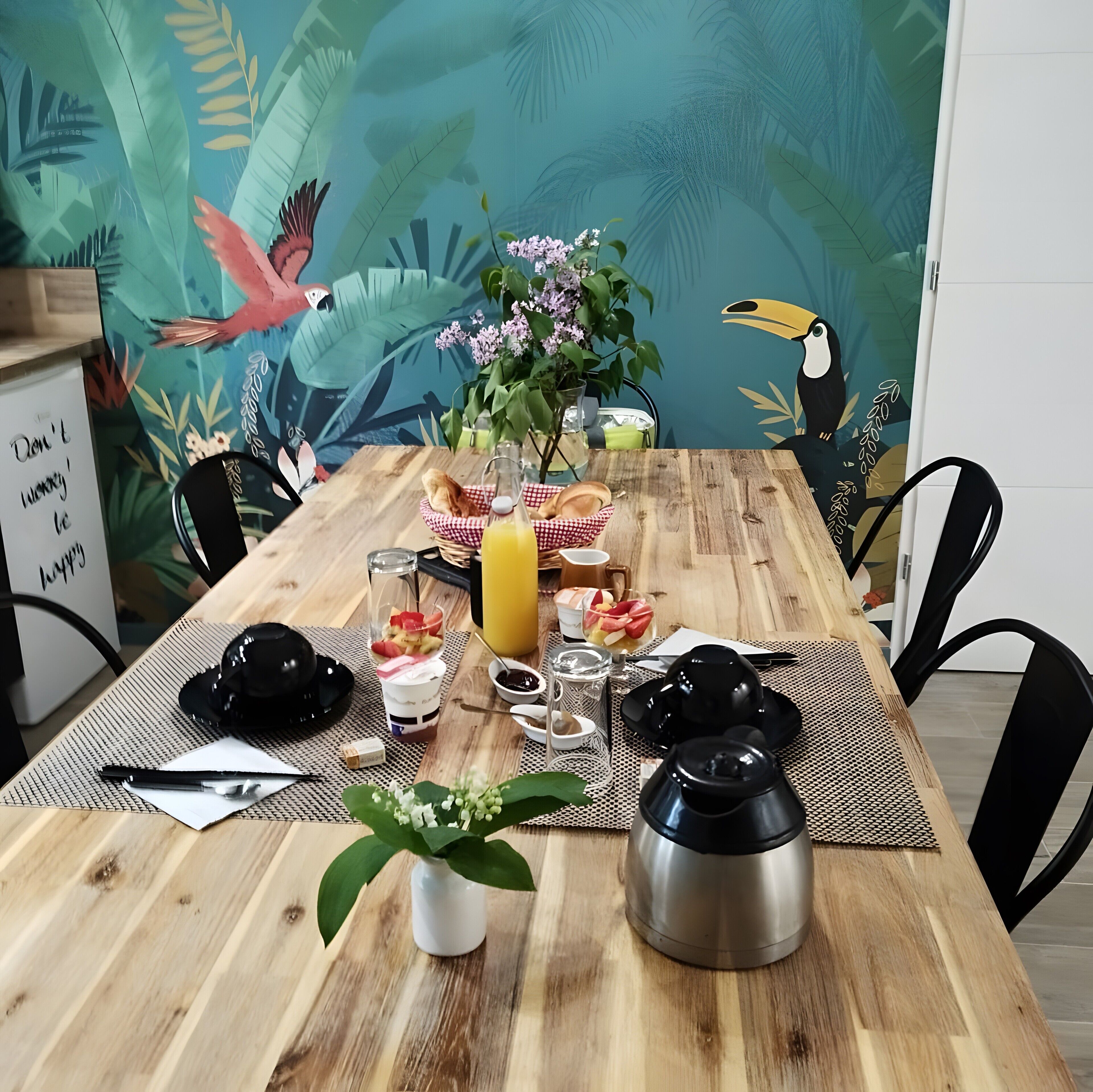 Ein Holztisch mit tropischem Wandbild, ein Glas Saft, eine Fruchtschale, ein Teekessel und eine kleine Pflanze.