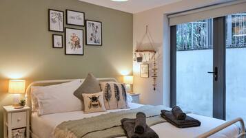 Quarto com cama, almofadas, mesa de cabeceira com abajur, decoração na parede e vista pela janela.