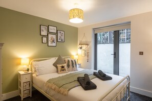 Une chambre à coucher avec un lit, des oreillers, une lampe de chevet, une lampe fixée au mur et des reproductions botaniques encadrées.