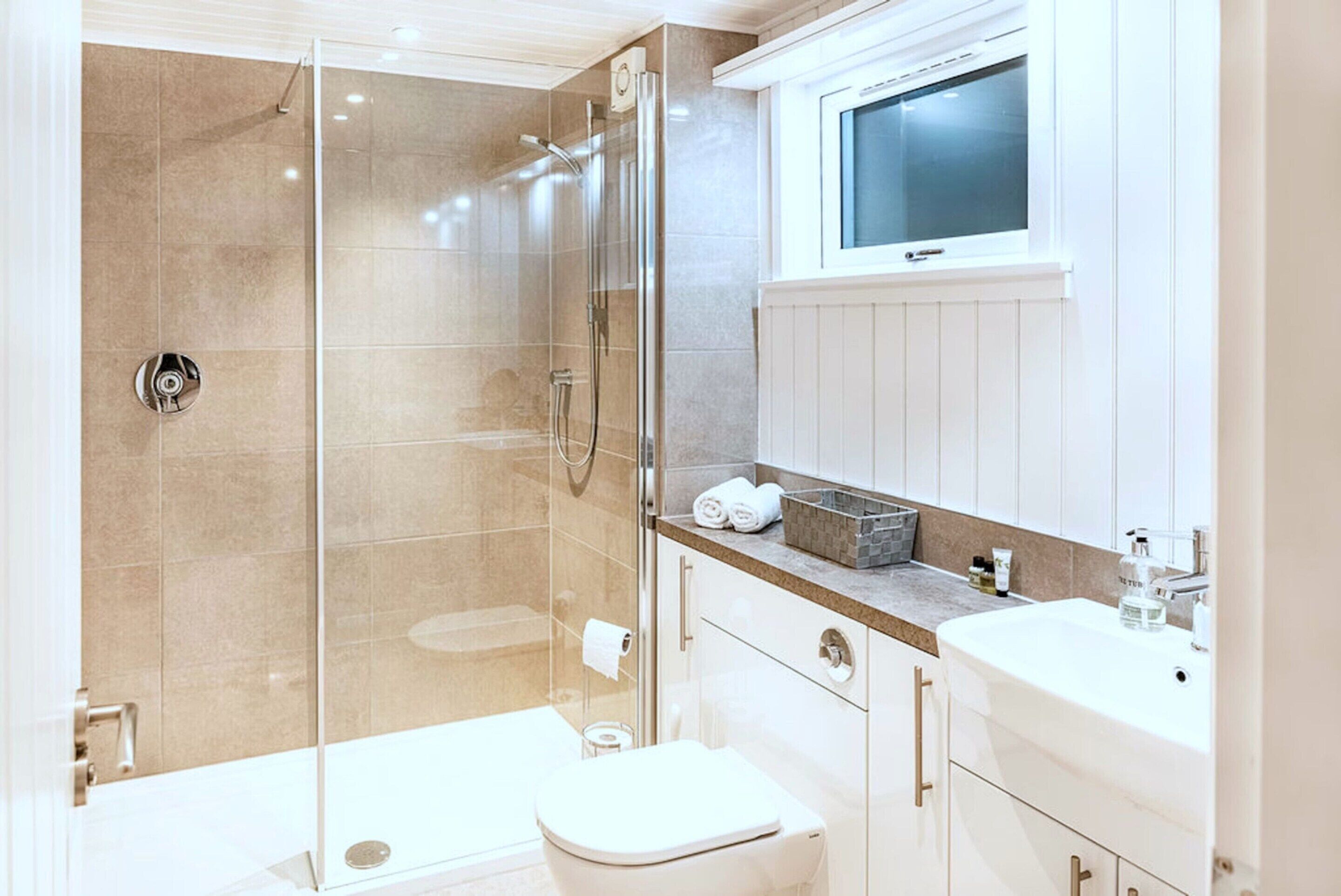 Un baño moderno con ducha de mampara de vidrio, inodoro, lavamanos y ventana sobre el espejo.