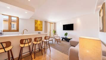 Una sala de estar moderna con área de cocina, taburetes de bar, un sofá y un televisor de pantalla plana montado en la pared.