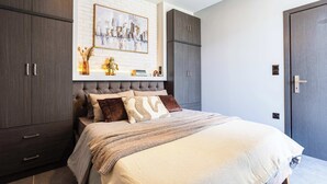 Un dormitorio con una cama grande, un armario de madera oscura, una pared de ladrillo blanco y un cuadro enmarcado con una vista urbana.