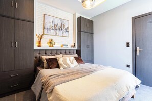 A bedroom with a large bed, dark wood wardrobe, a white brick wall, and a framed cityscape painting. - Socrates Suite C3 - Open City Views in Kalamata (Kalamata)