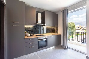 A modern kitchen with grey cabinets, a black oven, and a large window with a view of buildings and trees. - Socrates Suite C3 - Open City Views in Kalamata (Kalamata)
