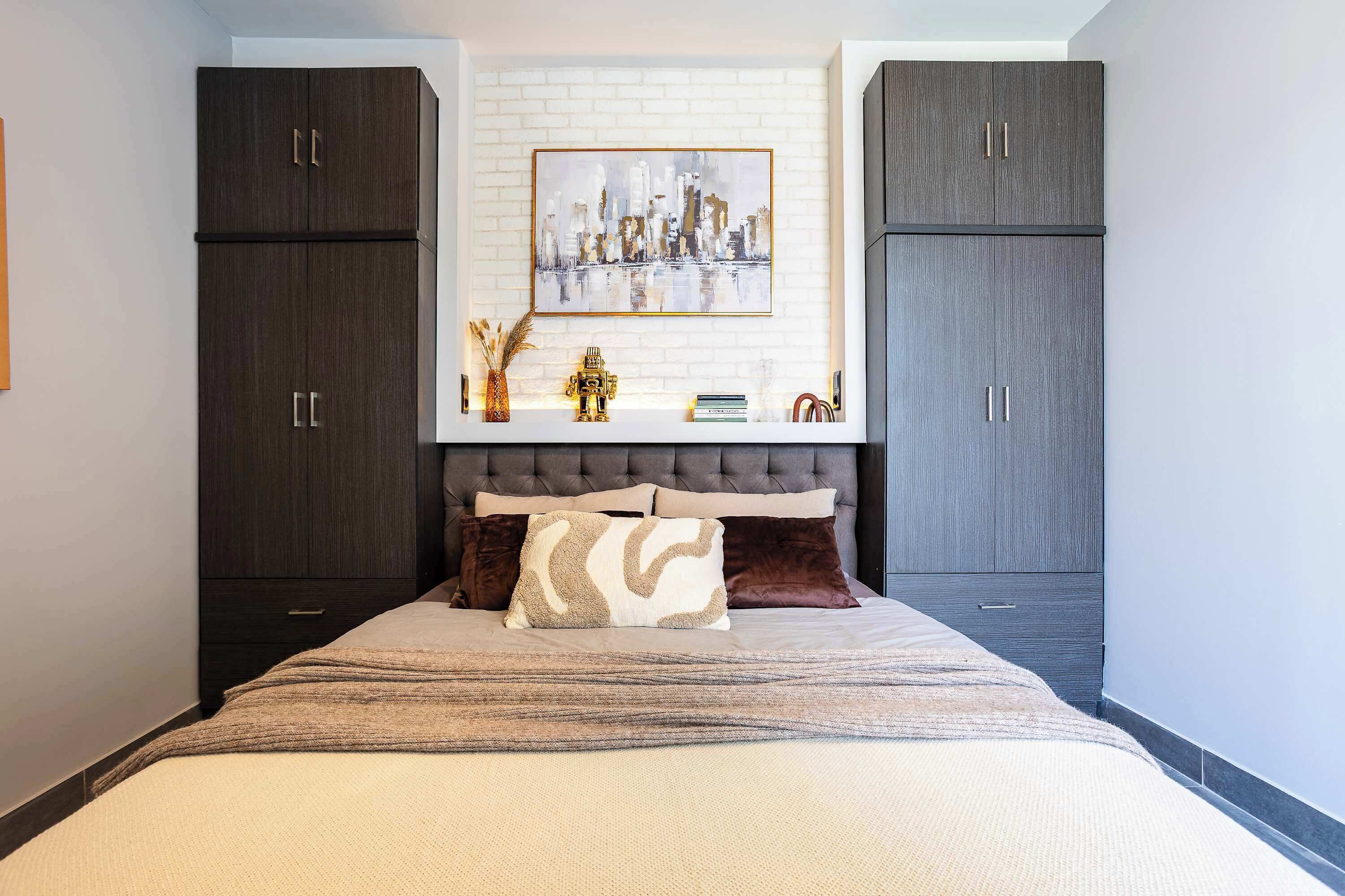 A bedroom with a bed, two dark wooden wardrobes, a cityscape painting, and a small decorative shelf with books and a golden figure.