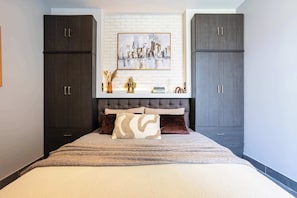 Ein Schlafzimmer mit einem Bett, zwei dunklen Holzschränken, einem Stadtbild als Gemälde und einer kleinen Dekorecke mit Büchern und einer goldenen Figur.