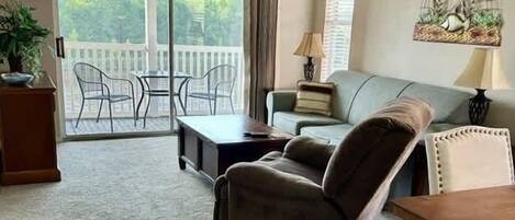 A living room with a ceiling fan, a sofa, a coffee table, and a TV. There is a balcony with a view of trees.