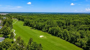 Ein Golfplatz unter blauem Himmel, umgeben von dichter Baumvegetation und mit Blick auf eine Stadt in der Ferne.