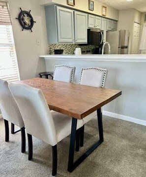 A dining area with a wooden table and white upholstered chairs, a kitchen in the background, and a carpeted floor. - Cozy Coastal Retreat With Scenic Waterway Views 16I (Myrtle Beach)