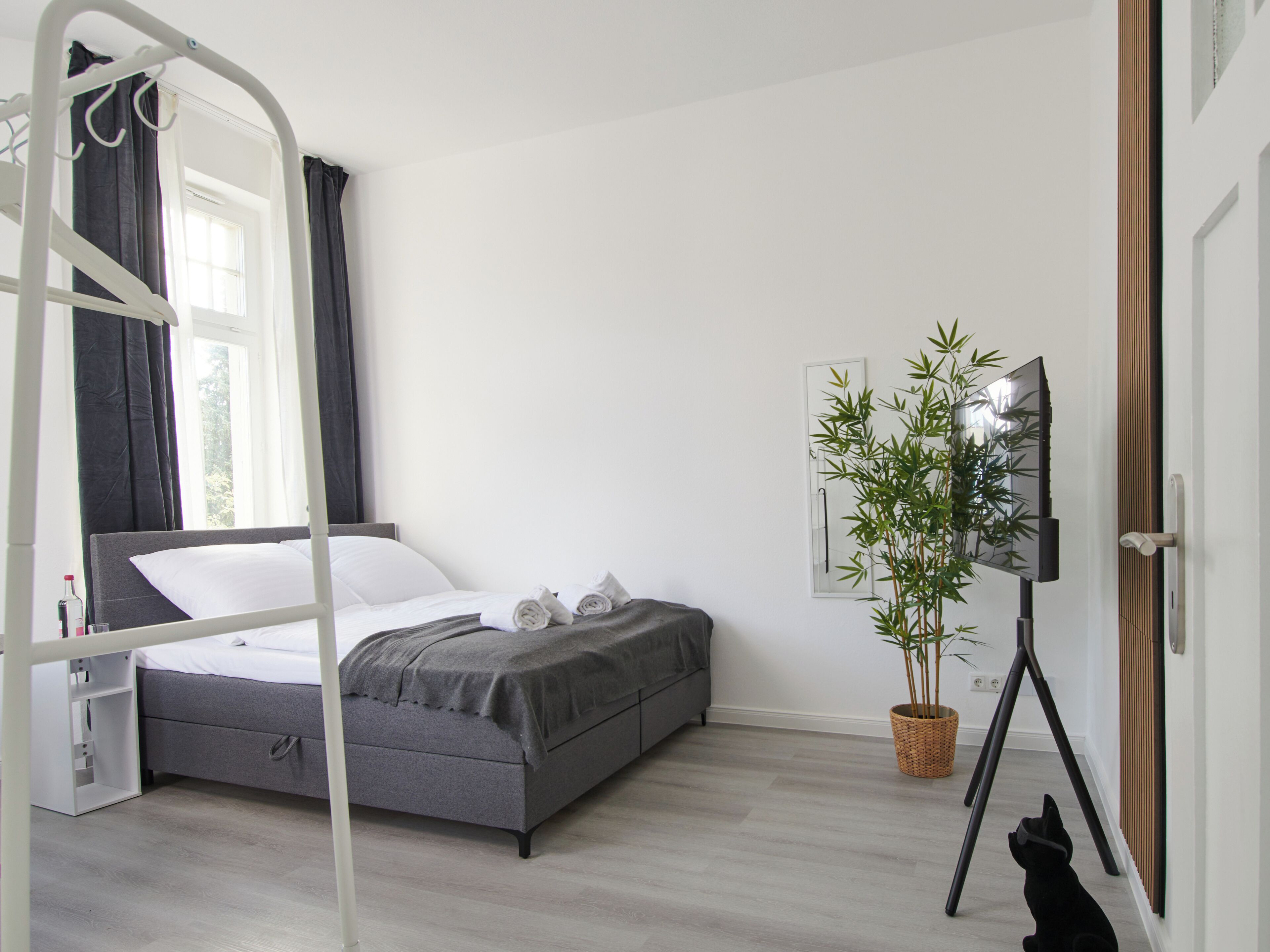 Een hotelkamer met een bed, een nachtkastje, een raam met gordijnen, een kamerplant en een televisie.