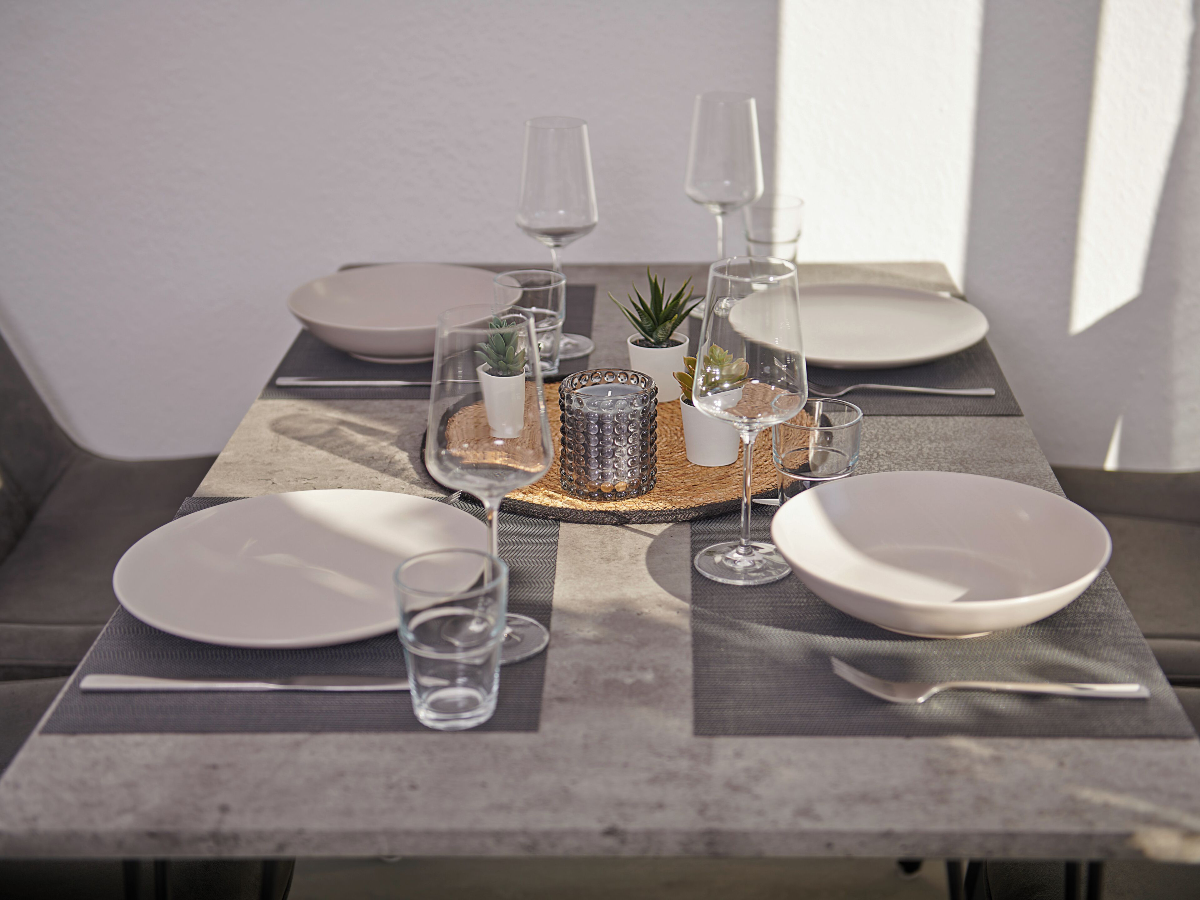 Una mesa de comedor dispuesta con platos blancos, vasos transparentes y una pequeña planta en maceta.