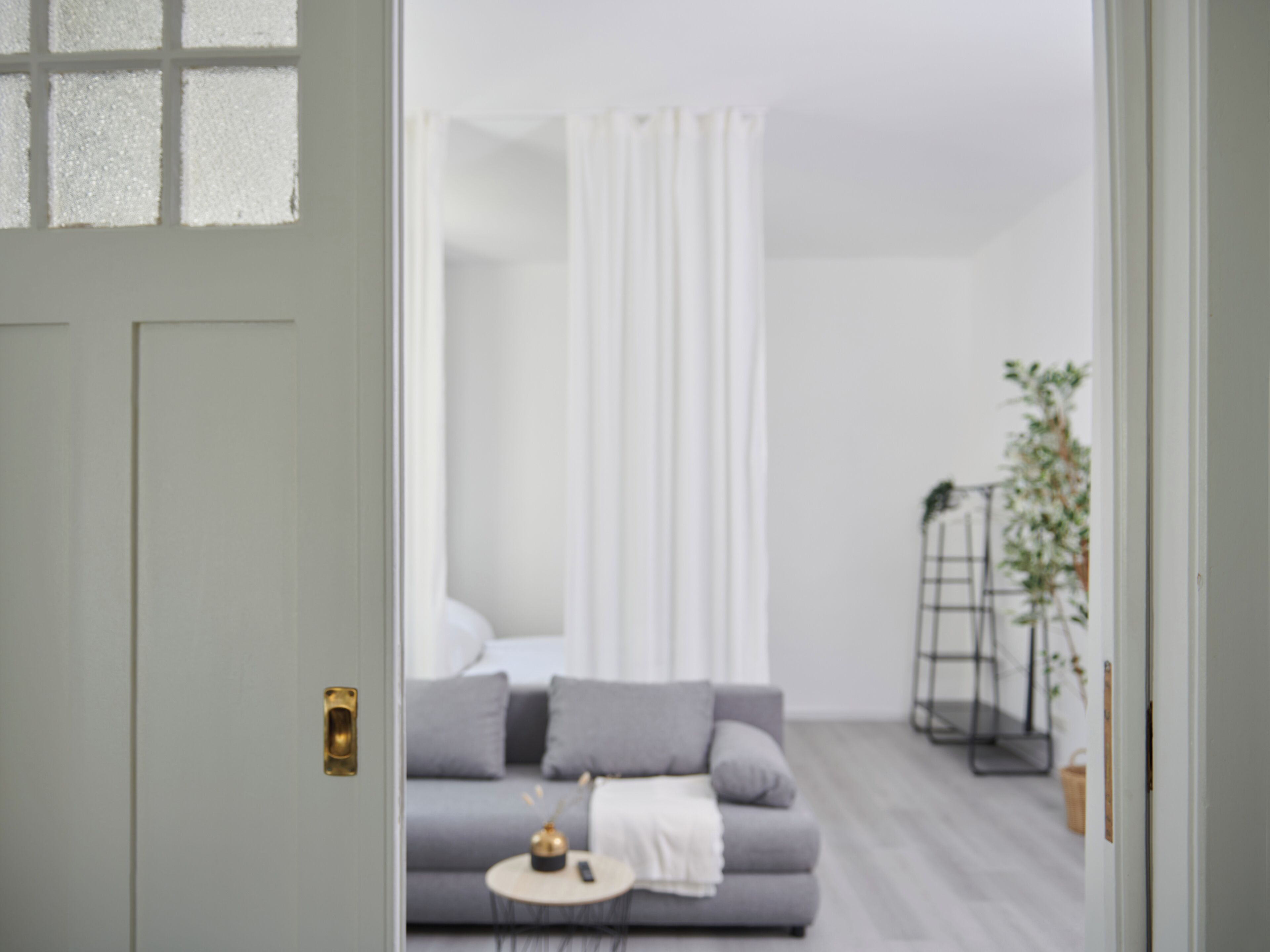 Ein modernes Wohnzimmer mit einem grauen Sofa, einem kleinen runden Tisch mit einer Fernbedienung und einem weißen Vorhang.