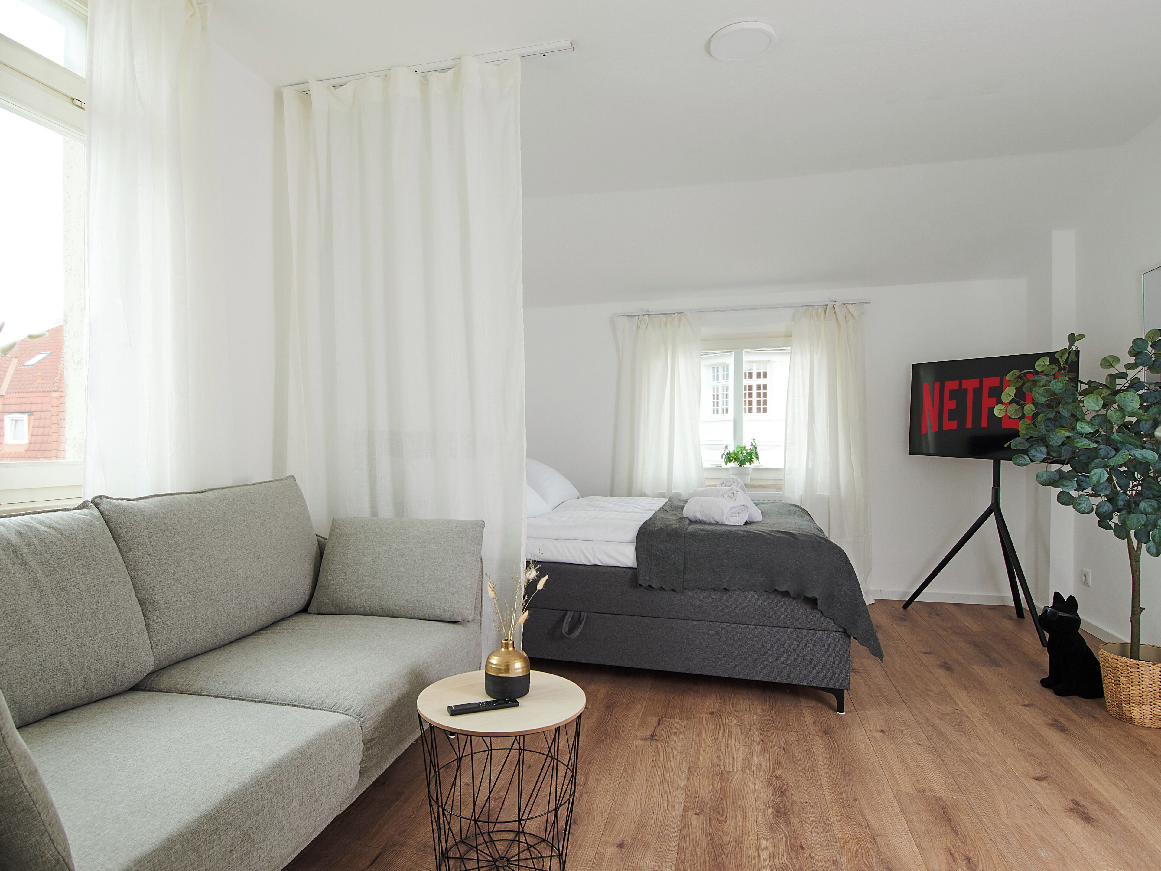 Um quarto moderno com um sofá cinzento, uma cama com roupa de cama cinza, uma mesinha redonda pequena com um vaso e um comando, e uma TV sobre um suporte de tripé.