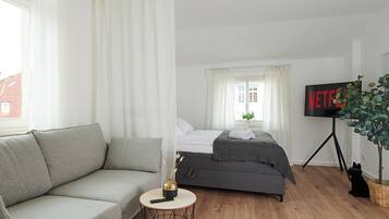 Um quarto moderno com um sofá cinzento, uma cama com roupa de cama cinza, uma mesinha redonda pequena com um vaso e um comando, e uma TV sobre um suporte de tripé.