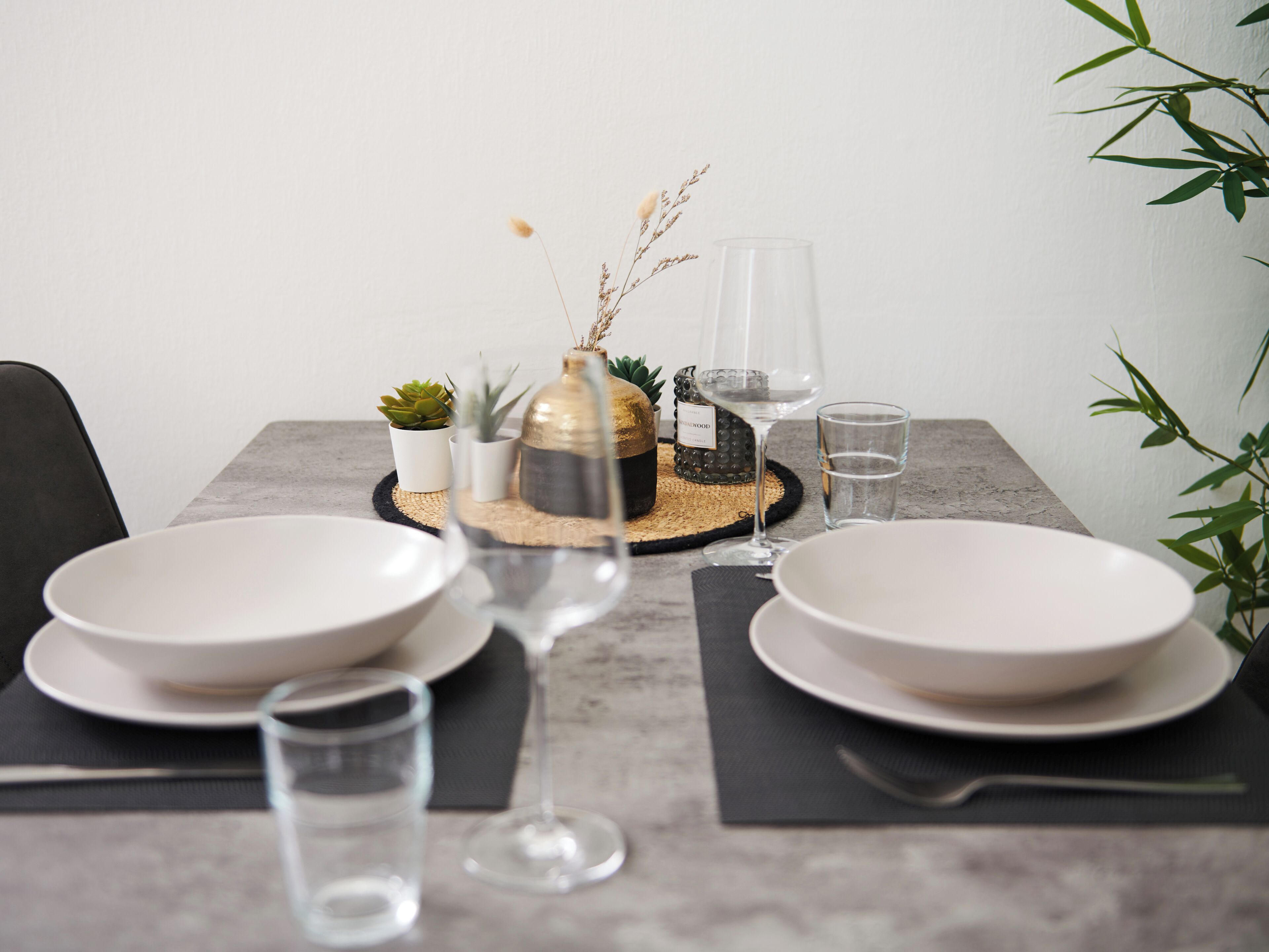 Een gedekte tafel met witte borden, een glas water, een wijnglas en een decoratieve vaas met gedroogde planten.
