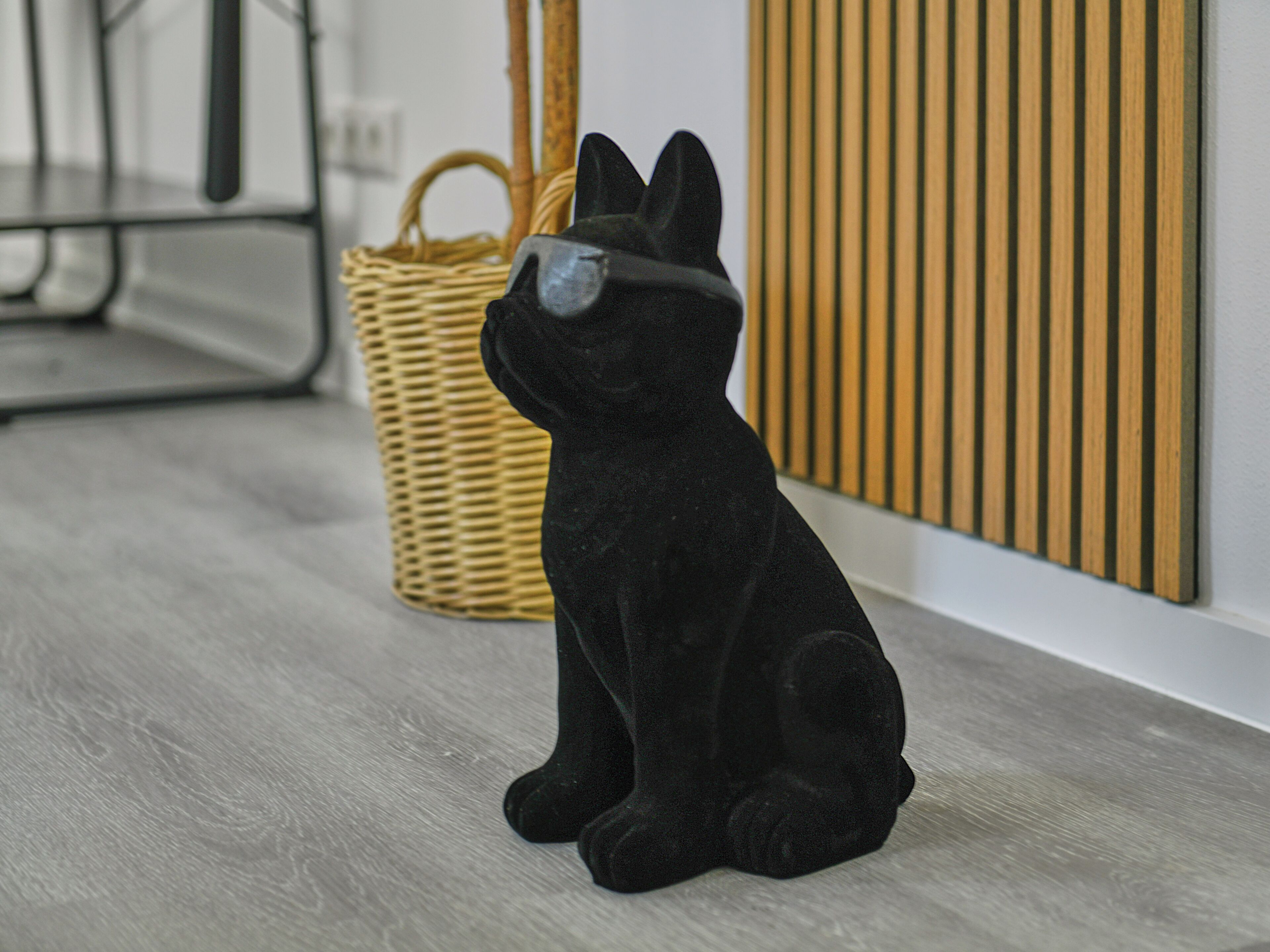 A black cat figurine on a wooden floor with a woven basket and a wooden panel in the background.