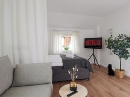 A modern hotel room with a grey sofa, a bed with white linens, a TV on a stand, and a potted plant.