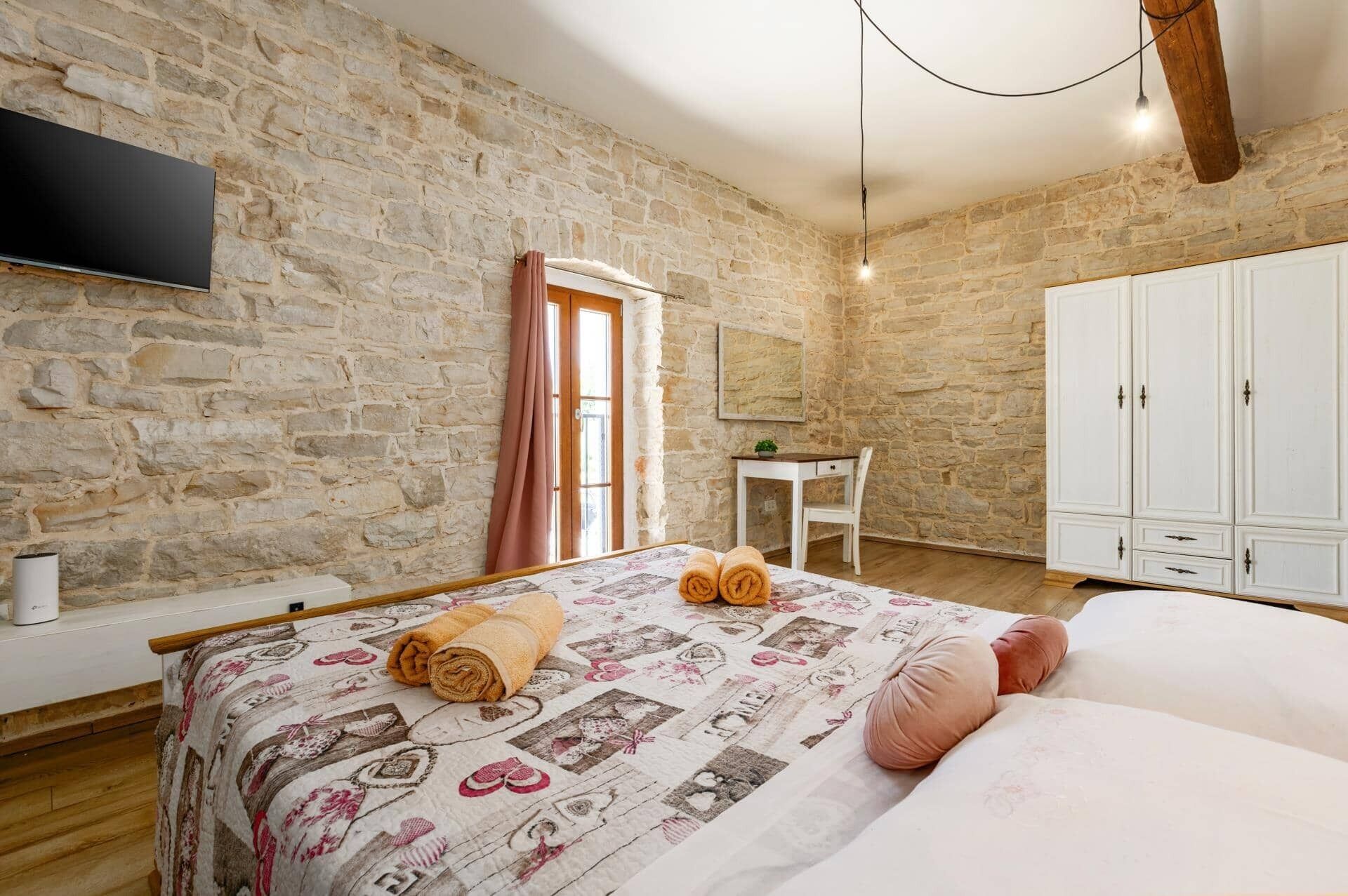 Une chambre avec un mur en pierre, un lit avec des serviettes roulées, un bureau et une armoire.