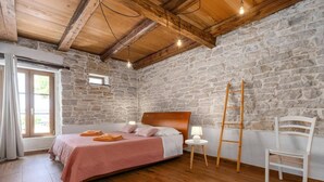Quarto com parede de pedra, vigas de madeira no teto, cama com roupa de cama rosa, luminária de cabeceira e uma cadeira branca.