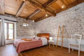 A bedroom with a stone wall, wooden ceiling beams, a bed with pink bedding, a bedside lamp, and a white chair. - Villa Lanca by Villa Plus (Heraki)