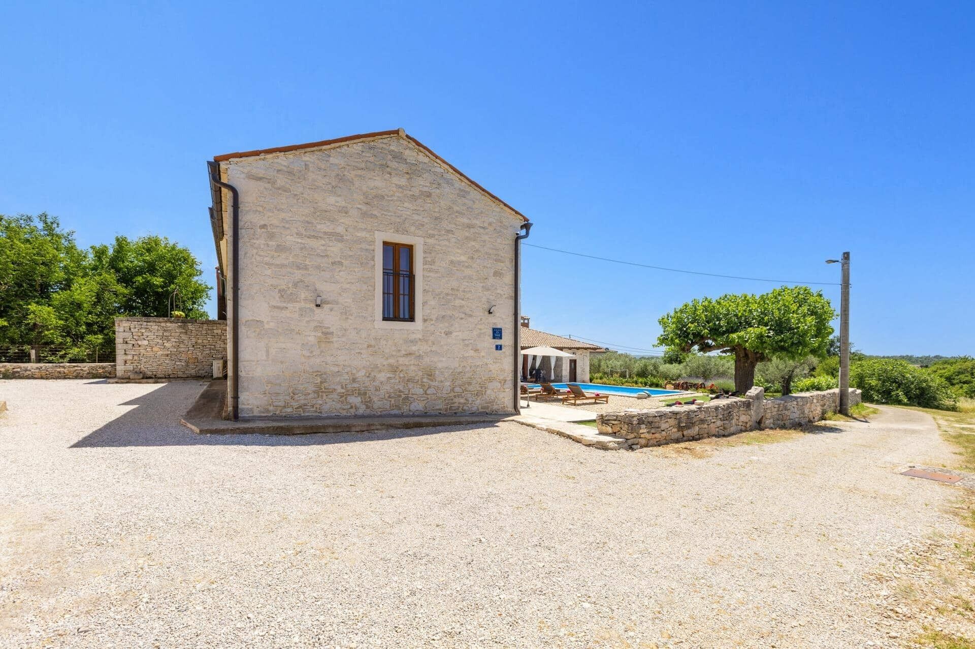 A stone building with a small window, a pool area with lounge chairs, and a tree in the background.