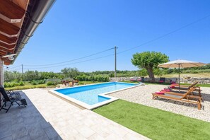 Une piscine bordée de pierres, entourée d’une terrasse aménagée avec du mobilier d’extérieur et un arbre.