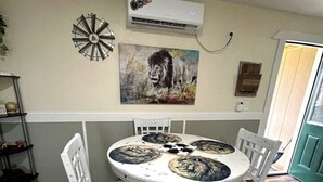 Zona de menjador amb una taula rodona, cadires blanques, aire condicionat fixat a la paret i un quadre amb un lleó.