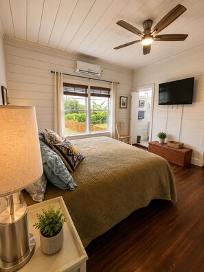 Ein Schlafzimmer mit einem Bett, einem Deckenventilator, einem Fernseher und einem Fenster mit Blick auf Grünflächen.