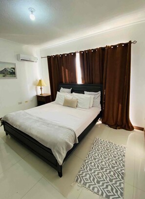 A hotel room with a bed, curtains, a nightstand with a lamp, and a patterned rug. - New Kingston Comfy 2BR Apartment  (Kingston)