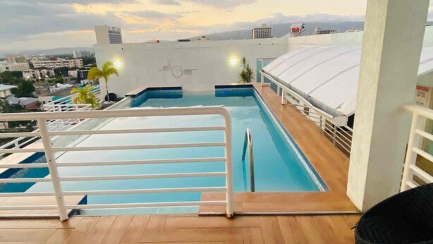 A rooftop pool with a wooden deck, surrounded by white railings, and overlooking a cityscape. - New Kingston Comfy 2BR Apartment  (Kingston)