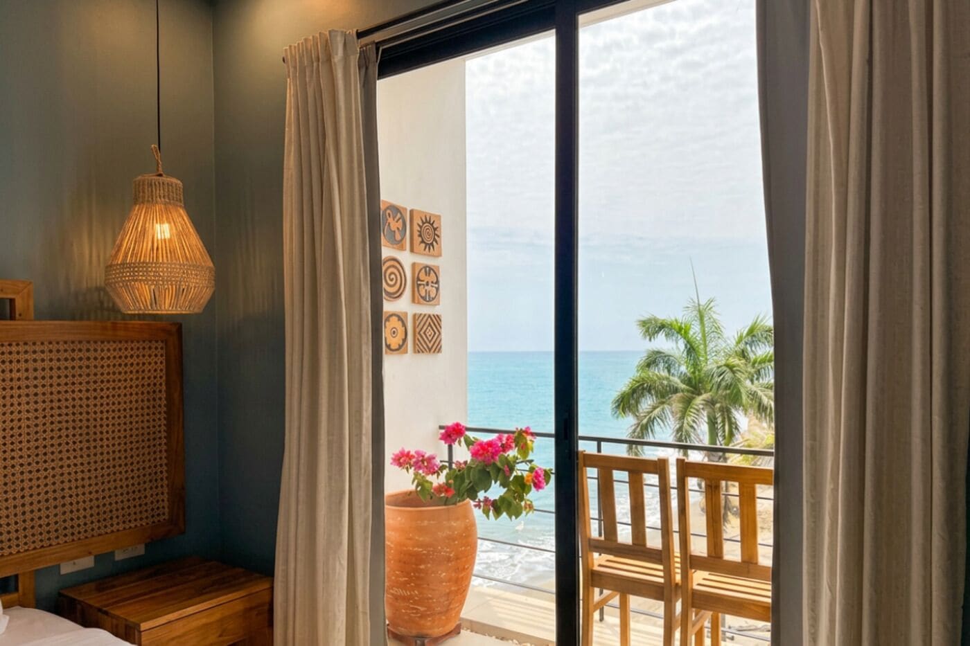 A room with a bed, a woven headboard, a hanging light fixture, a large potted plant, and a balcony with a view of the sea and palm trees.
