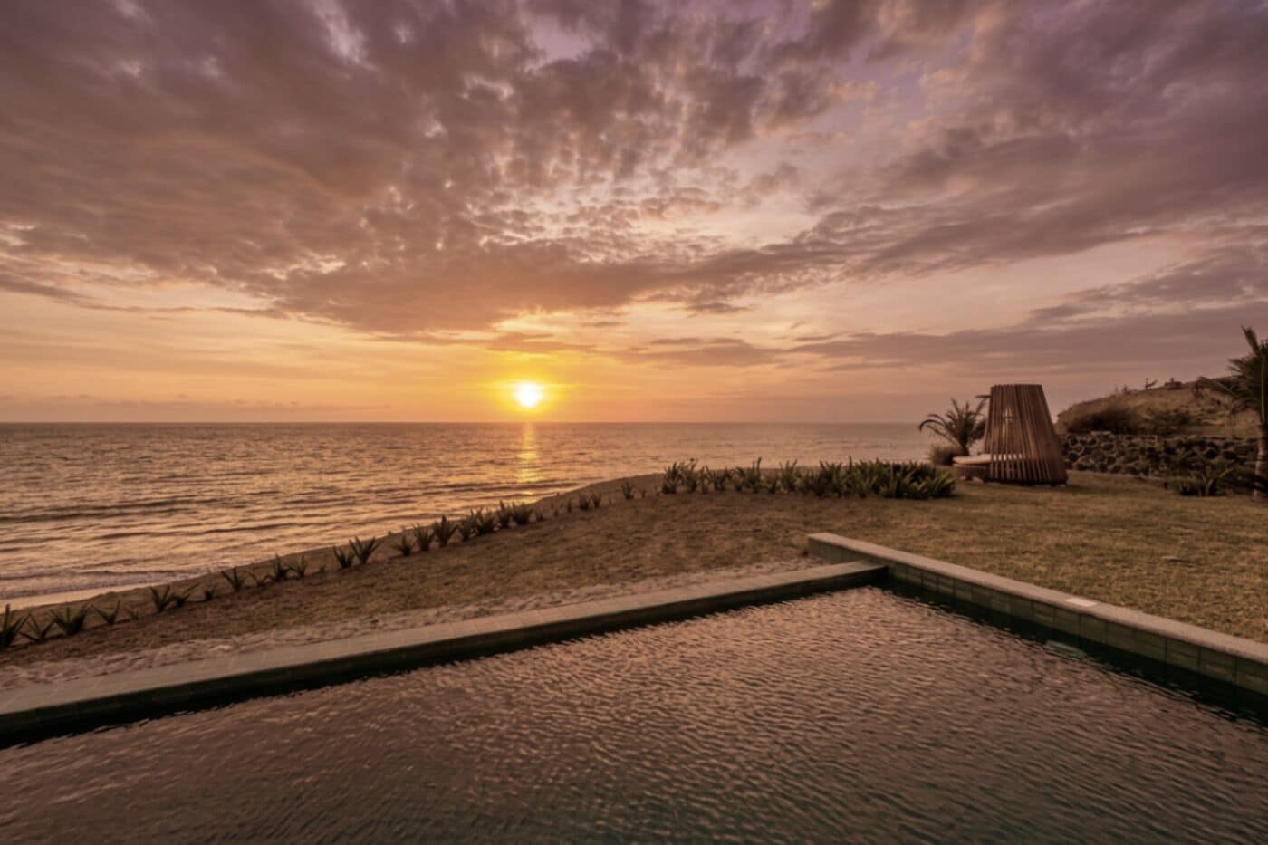 A pool area with a view of the sunset over the ocean, a lounge chair, and a small garden.