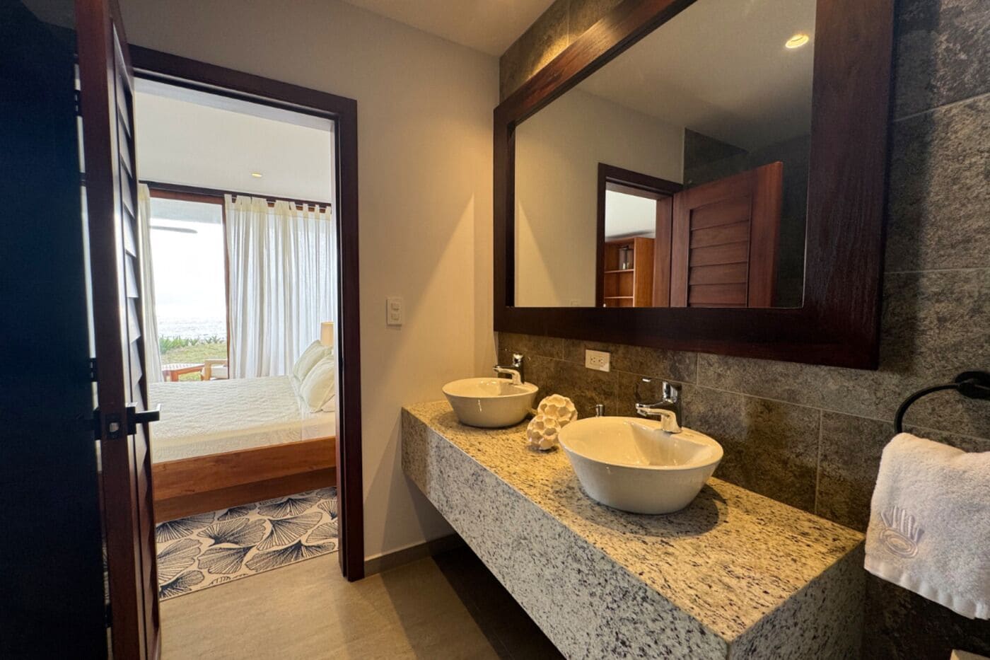 A bathroom with two sinks, a large mirror, and a view of a bedroom with a bed and curtains.