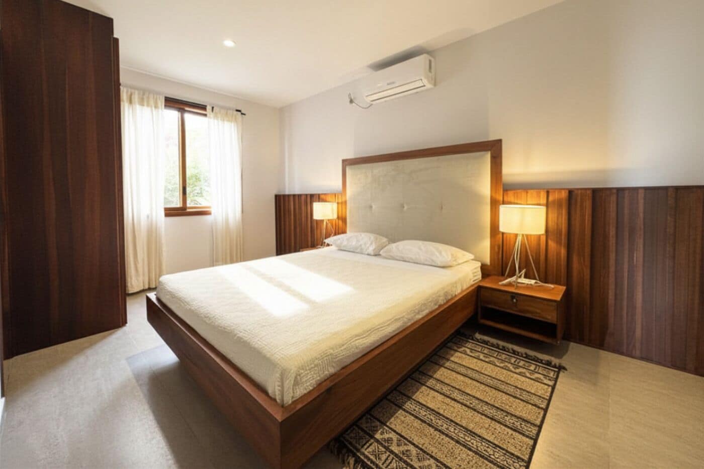 Quarto de hotel com cama grande, luminárias de cabeceira, ar condicionado e janela com cortinas.