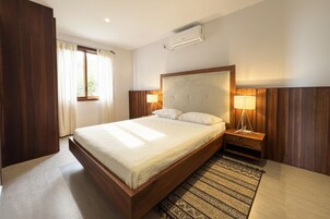 A hotel room with a large bed, bedside lamps, an air conditioning unit, and a window with curtains.