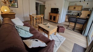A living room with a brown sofa, a dining table with chairs, a TV on a wooden cabinet, and a kitchen area with wooden cabinets and a bar counter. - APARTMENT NEAR LUZ SAINT SAUVEUR 4/5 -PERSON APARTMENT (Esquièze-Sère)