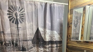 A bathroom with a shower curtain depicting a windmill and a cow, a sink, and wooden cabinets. - Western cabin in Blanchard, OK (Blanchard)