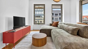 A modern living room with a flat-screen TV on a red cabinet, a grey sofa with brown cushions, a wooden coffee table, and a large window with a view of buildings. - Scandi & Modern Apartment in Heart of Bratislava (Bratislava I)