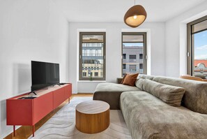 A modern living room with a flat-screen TV on a red cabinet, a grey sofa with brown cushions, a wooden coffee table, and a large window with a view of buildings. - Scandi & Modern Apartment in Heart of Bratislava (Bratislava I)