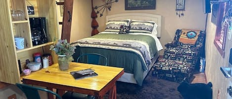 A cozy room with a bed, a table with chairs, a shelf with various items, and a small rug.