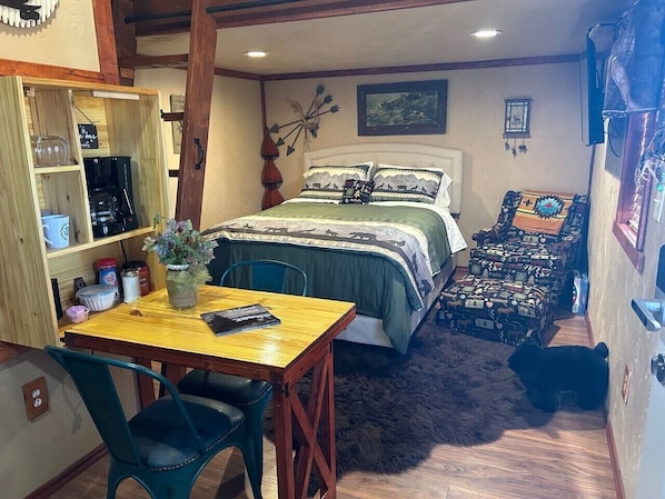 A cozy room with a bed, a table with chairs, a shelf with various items, and a small rug.