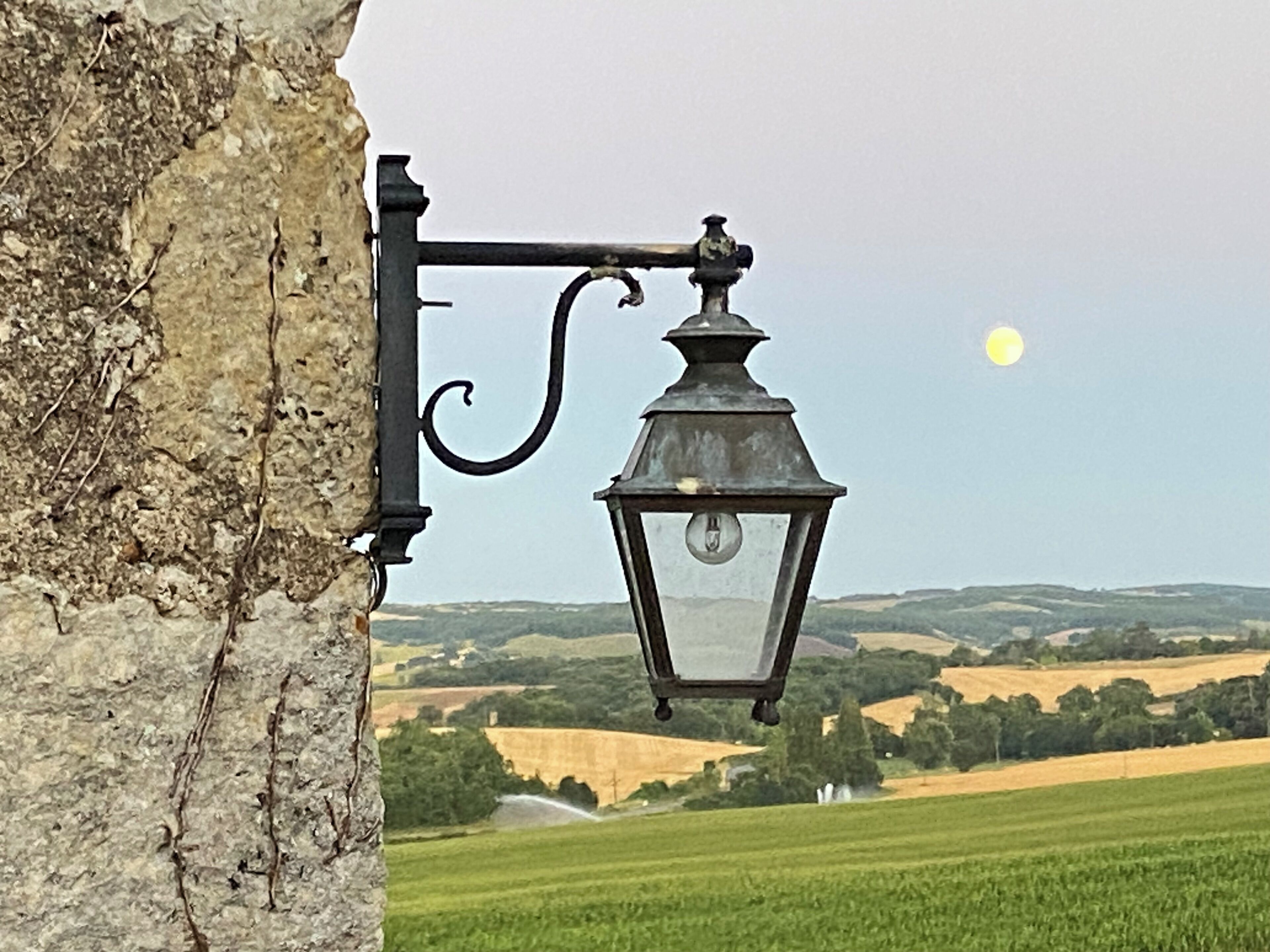 Un lampadaire fixé à un mur de pierre, surplombant un paysage rural avec des champs et des collines lointaines.