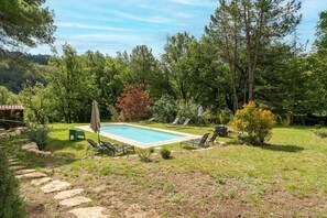 Ein rechteckiger Pool mit Liegestühlen und einer kleinen Laube in einem üppigen, bewaldeten Garten.
