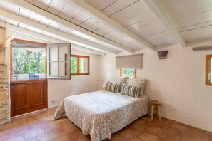 Ein Schlafzimmer mit einem Bett, einem hölzernen Hocker, einem Fenster mit Blick auf Bäume und einer Steinsäule.