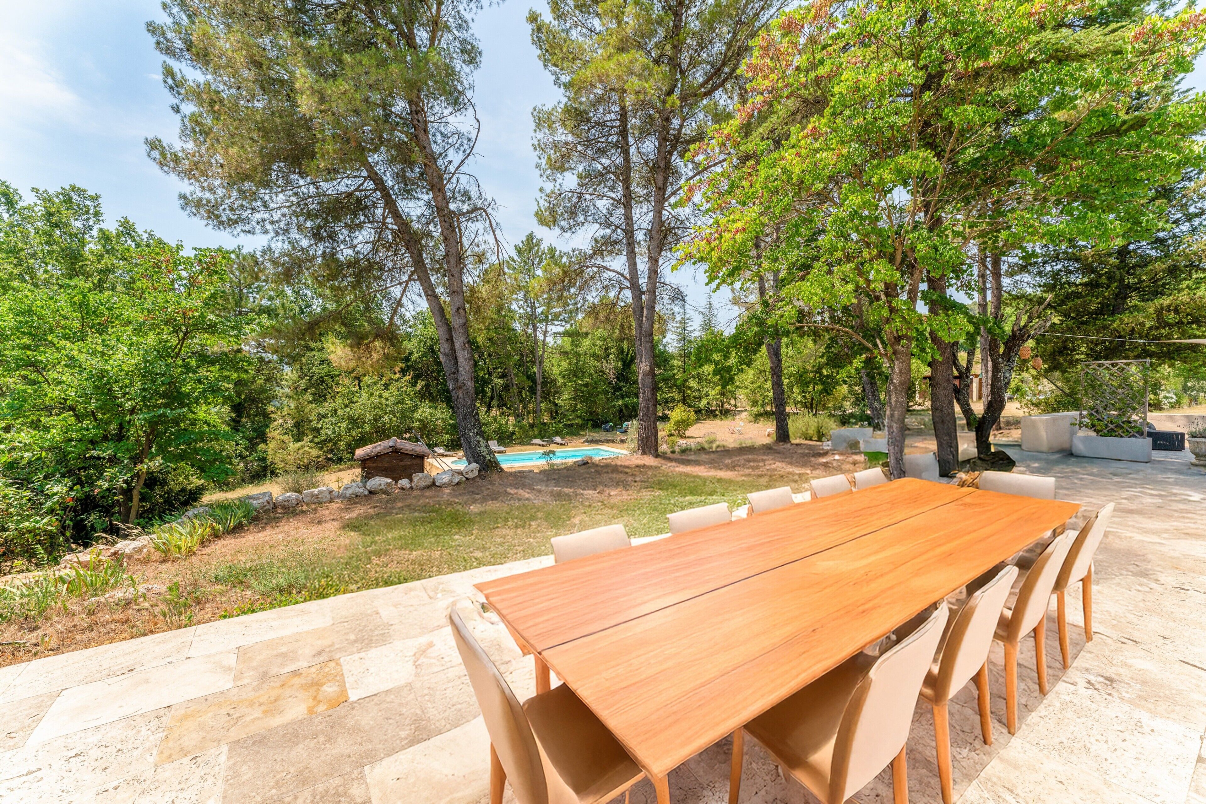 An outdoor dining area with a long wooden table and chairs, surrounded by trees and a swimming pool.