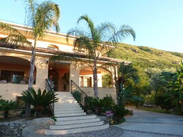 Un edificio de dos pisos con una escalera que conduce a la entrada, rodeado de palmeras y un muro de piedra.
