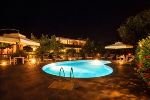 A nighttime swimming pool area with lounge chairs and umbrellas, surrounded by a building with illuminated windows. - Hotiday Tropea Parghelia (Parghelia)